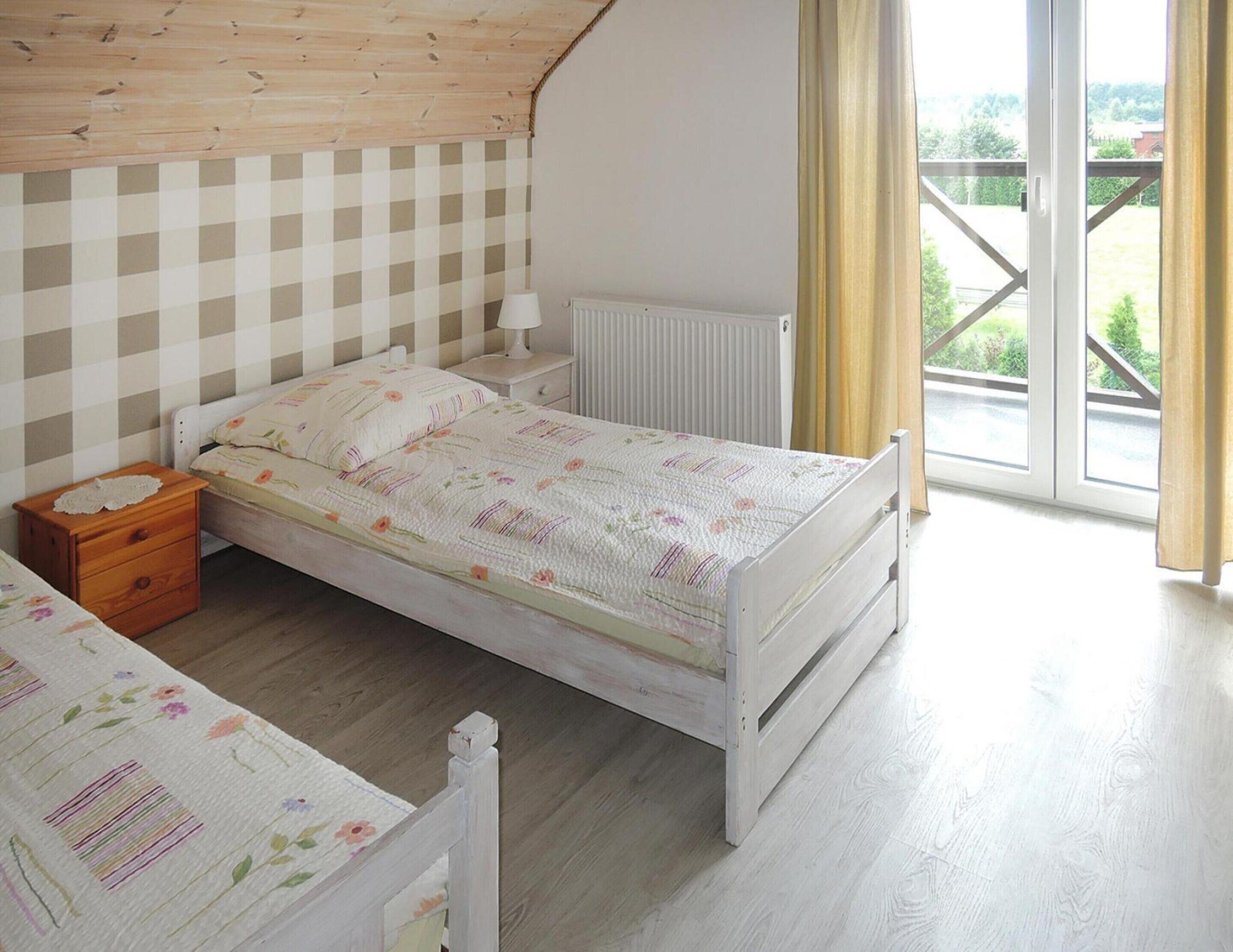 Ein Schlafzimmer mit Holzboden, einer karierten Wand, einem Bett mit floralem Bettzeug, einem Nachttisch, einem Heizkörper und Blick ins Grüne durch ein Fenster.