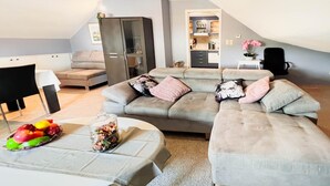 Ein modernes Wohnzimmer mit einem grauen Ecksofa, einem Esstisch mit Obstschale und einer Küche im Hintergrund.