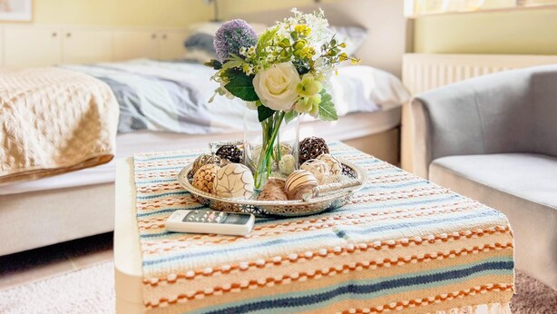 Ein gemütliches Zimmer mit einem Bett, einem grauen Sofa, einem kleinen Tisch mit Blumen und einer Platte Kekse.