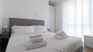 A modern bedroom with a large bed, white linens, and a grey headboard. There are two bedside tables with lamps and a stack of folded towels on the bed. - Italianway Comfort - Boscovich 23 (Milan)