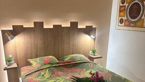 Una cama bien tendida con una colcha estampada, una mesita de noche con una planta y una lámpara de pared sobre la cama.