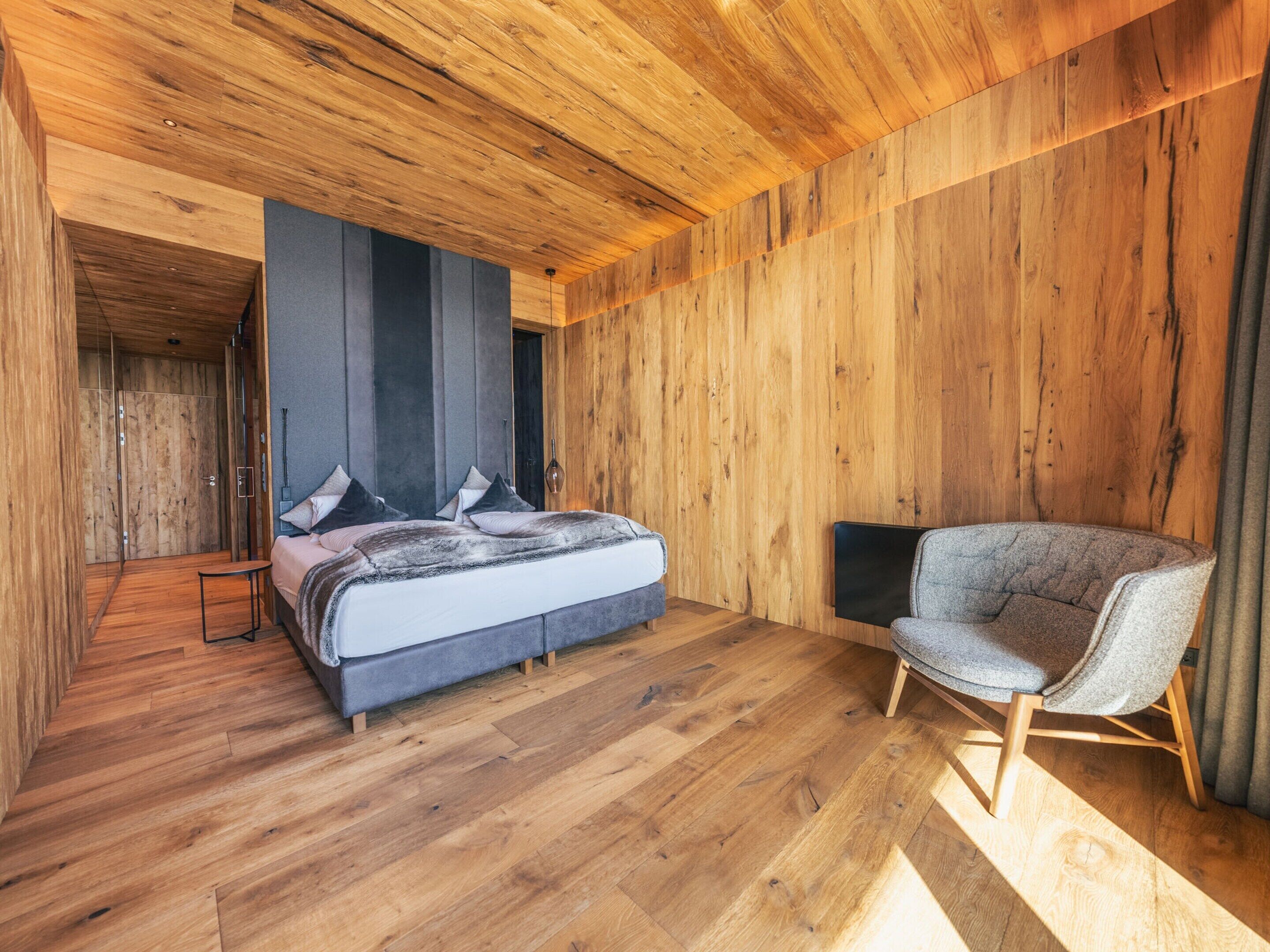 A modern bedroom with a wooden ceiling, a bed with grey bedding, a grey armchair, and a wooden wardrobe.
