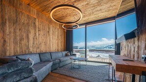 Ett modernt vardagsrum med ett träpanelat tak, ett stort fönster med utsikt över bergen, en grå hörnsoffa och ett litet bord med stolar.