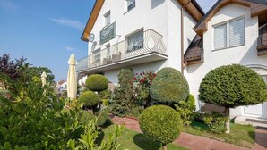 Una casa de dos pisos con un balcón, rodeada de jardines cuidados y un sendero pavimentado.