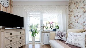 Una sala de estar acogedora con una televisión de pantalla plana, un sofá con almohadones decorativos, un aparador de madera y un amplio ventanal con cortinas.