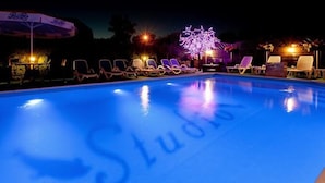 Piscina con sillones de descanso y sombrillas, rodeada de árboles y un edificio con ventanas iluminadas.