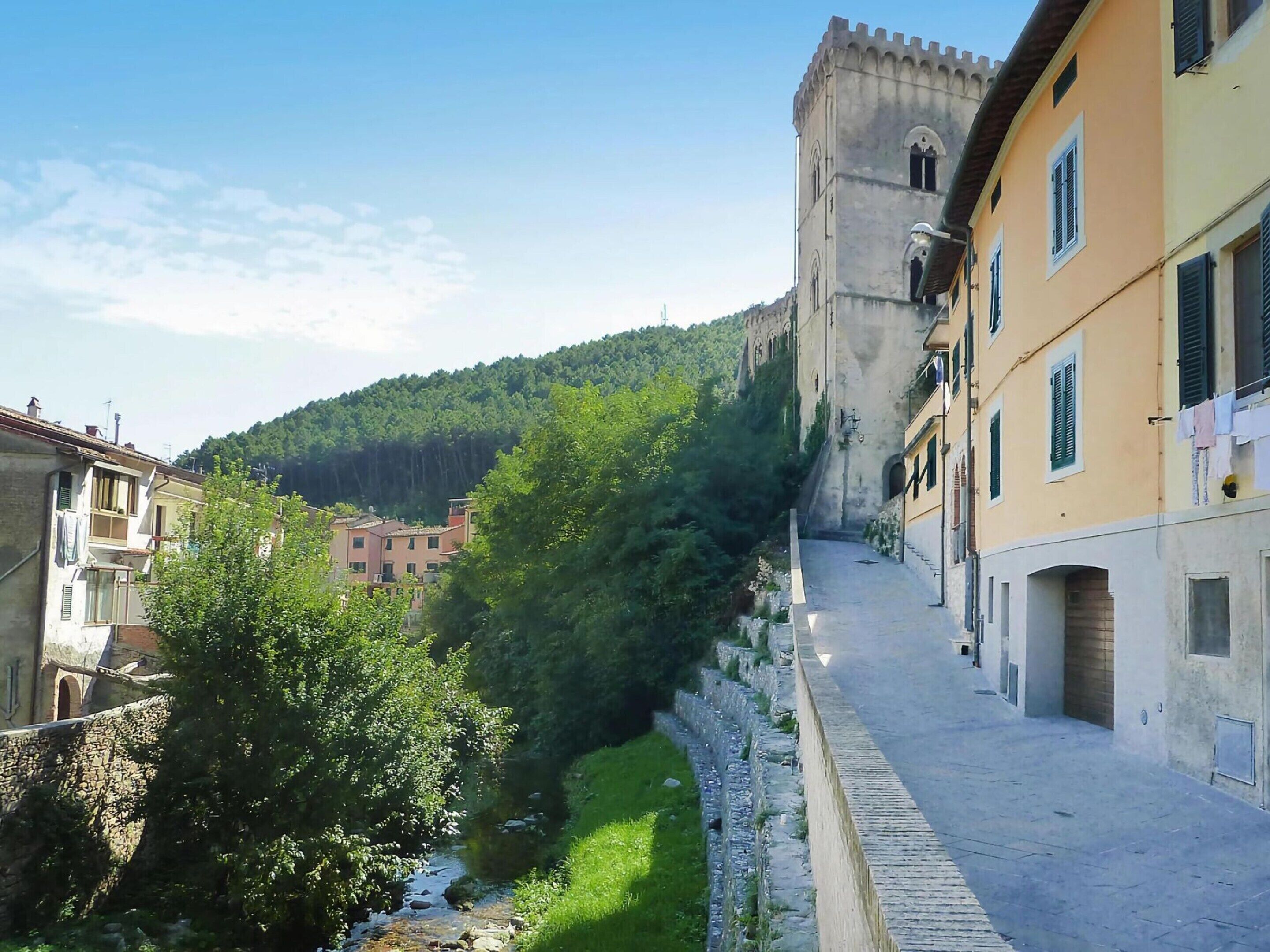 A narrow street with stone walls and buildings on either side, leading to a hill with a castle-like structure.