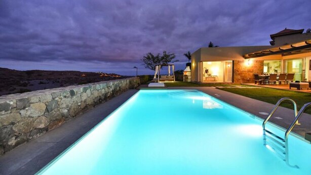 A swimming pool with a stone wall on one side and a building with outdoor seating on the other. - Holiday Home With Pool on Golf Resort, Maspalomas (San Bartolomé de Tirajana)