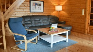 A cozy cabin interior with a wooden staircase, a leather sofa, a blue armchair, a small white table with a fruit bowl, and a framed abstract painting on the wall.