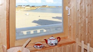 A wooden cabin with a window overlooking a beach. Inside, there is a wooden table with a teapot and cup. - Doppelhaushalfte, Ostseeresort Olpenitz (Kappeln)