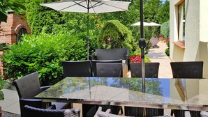 Eine Terrassengruppe im Freien mit einem Glastisch und Korbstühlen, umgeben von üppiger Bepflanzung und einer Ziegelstruktur.