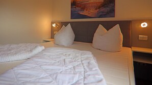 A hotel room with a large bed, two pillows, a nightstand, a wall-mounted lamp, and a painting of a sunset over water. - Appartementhaus Nicole, Zinnowitz (Zinnowitz)