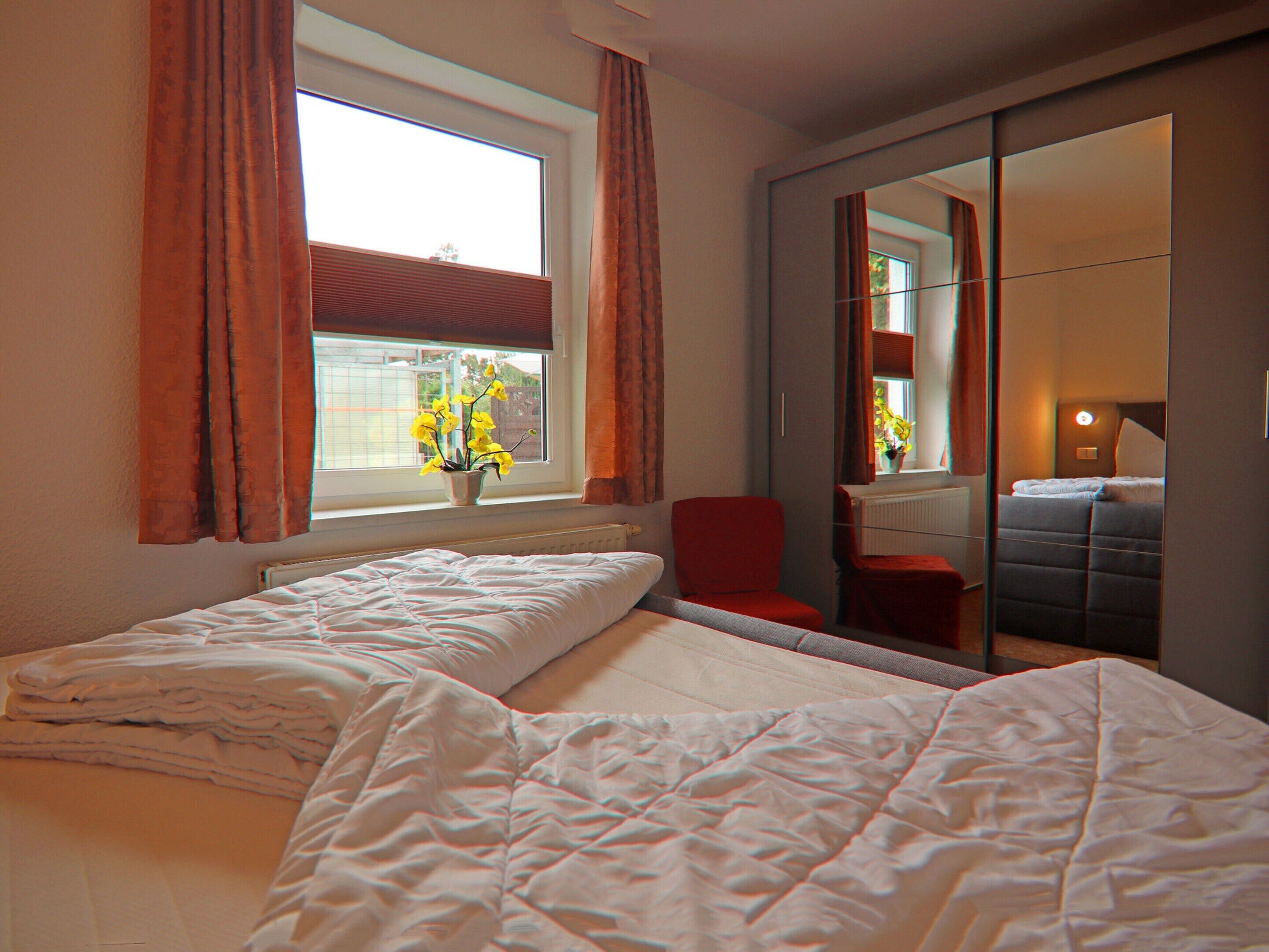 Ein Hotelzimmer mit zwei Betten, einem mit Vorhängen versehenen Fenster, einem roten Sessel, einer Zimmerpflanze und einem Spiegel, der den Raum widerspiegelt.