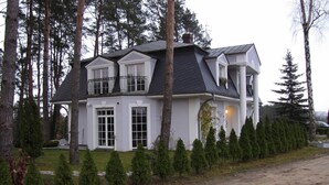 Une maison à deux étages avec un balcon, entourée d’arbres et donnant sur une allée gravillonnée.