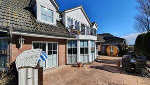 A two-story house with a large patio, a round wooden structure, and a white wicker chair. - Charmante Hausvilla in Bojendorf (Fehmarn)