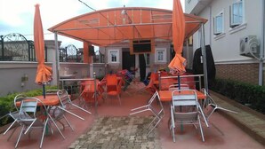 An outdoor patio area with metal chairs and orange umbrellas, a brick pathway, and a building in the background. - Prodis Hotel and Suites (Sagamu)