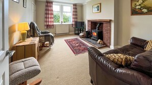 A cozy living room with a fireplace, a sofa, a chair, a small table with a lamp, and a window with curtains. - 2 bedroom accommodation in Castle Douglas (Castle Douglas)