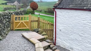 A stone pathway leading to a wooden gate, a white building with a red pipe, and a stone wall with a spherical tree. - 2 bedroom accommodation in Castle Douglas (Castle Douglas)