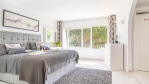 Ein modernes Schlafzimmer mit einem großen Bett, einem gerahmten Kunstwerk darüber und Blick ins Freie durch große Fenster.
