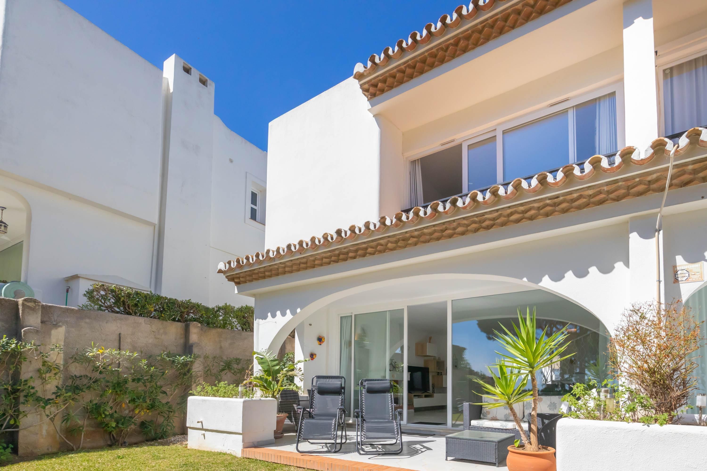 A two-story building with a white exterior, a balcony with chairs, and a glass door leading to a patio area with plants.