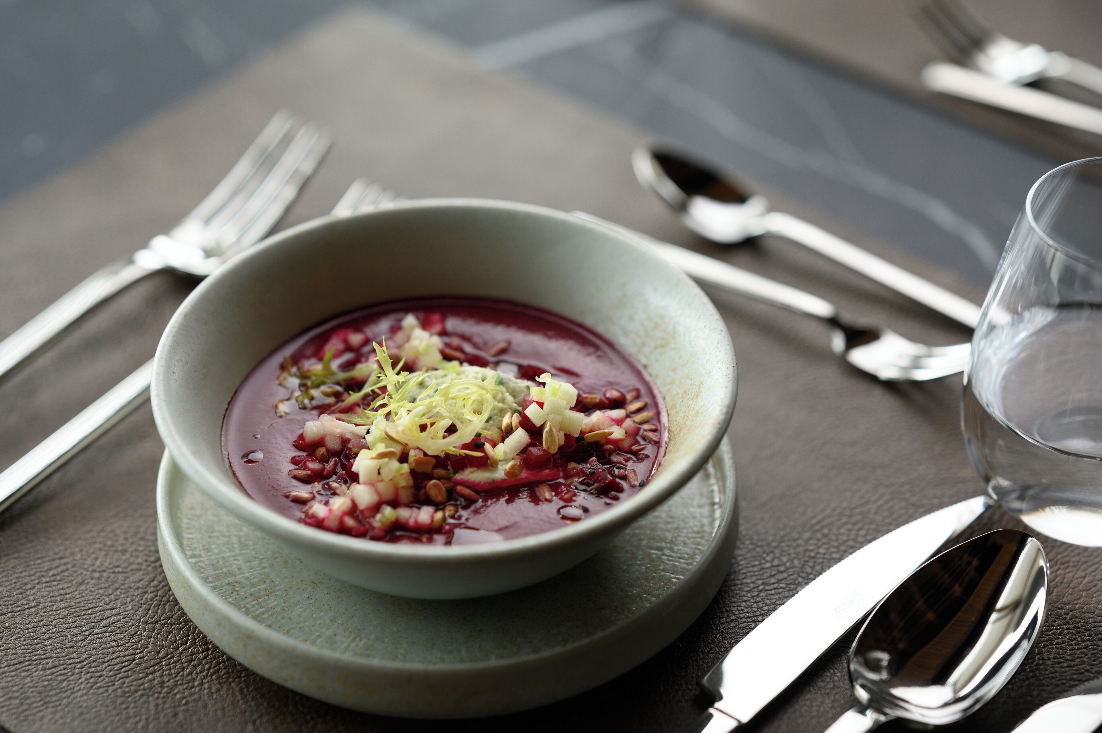 A bowl of soup with garnishes on a plate, accompanied by cutlery and a glass of water on a table.
