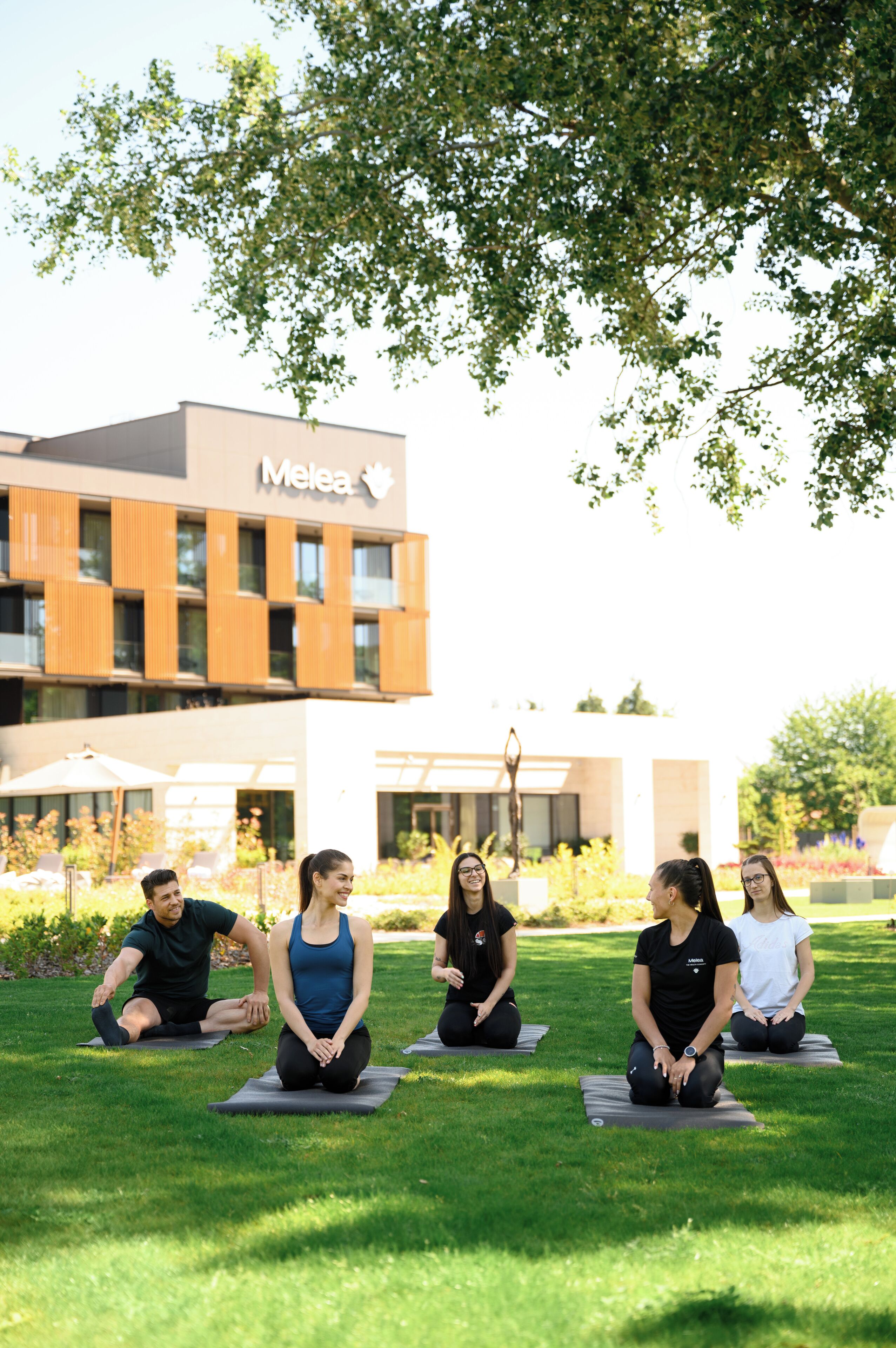 Un groupe de personnes est assis sur des tapis de yoga dans un espace verdoyant, avec un bâtiment en arrière-plan.