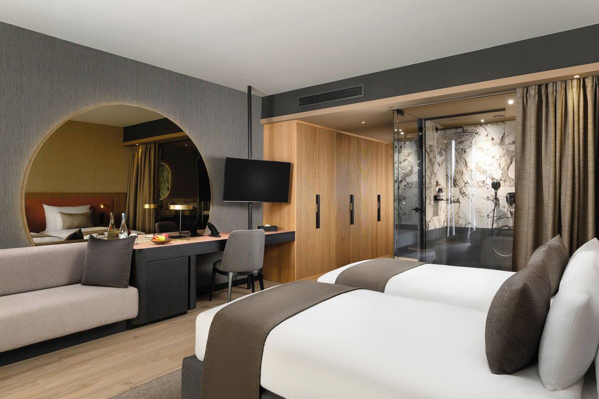 A modern hotel room with a sofa, two beds, a desk, and a television.