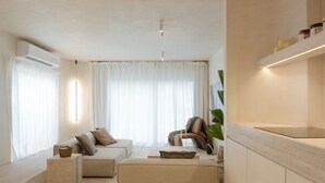 En moderne stue med sofa, skammel og sofabord. Der er et stort vindue med lette gardiner, en aircondition og en plante.