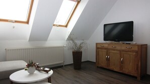 Ein Zimmer mit schrägverdeckter Decke, einem weißen Heizkörper, einem Holzschrank, einem Fernseher, einem kleinen weißen Tisch und einer Pflanze.