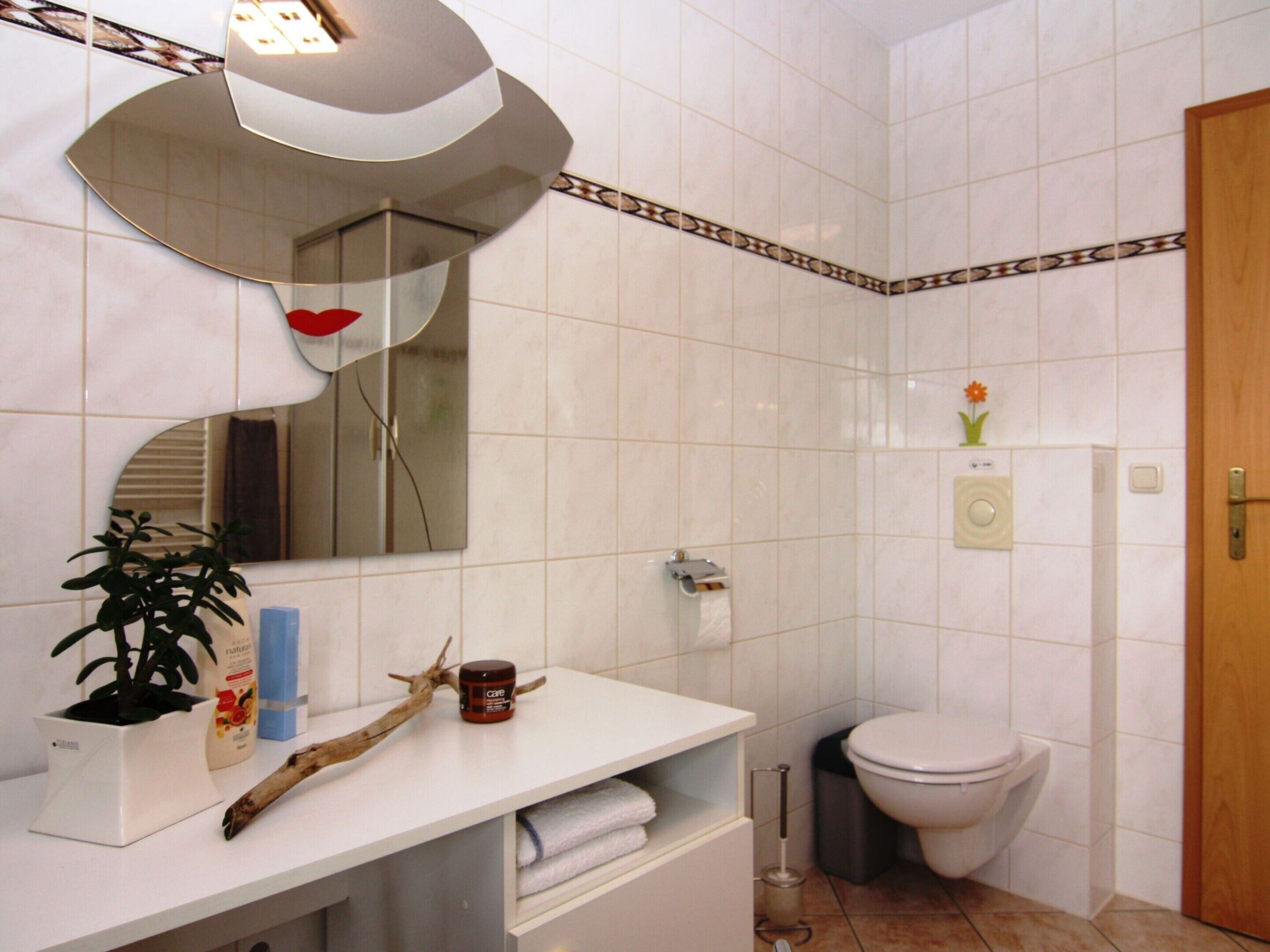 Ein Badezimmer mit weißen Fliesen, einem weißen Waschbecken, einem Spiegel, einer Toilette und einer kleinen Pflanze.