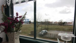 Ein Zimmer mit einem großen Fenster, einer weißen Vase mit rosa Blumen und einem kleinen Tisch mit einer Kerze.