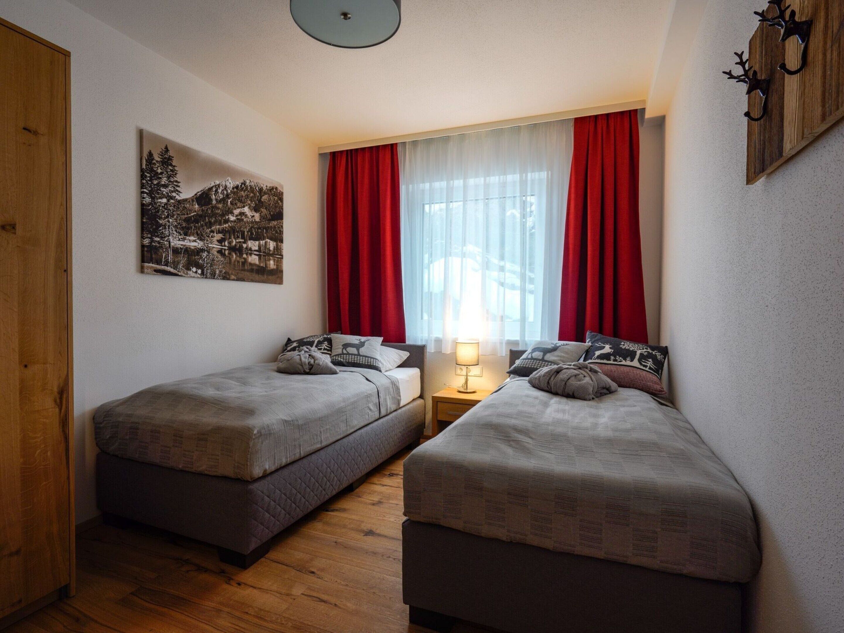 Ein kleiner, gemütlicher Raum mit zwei Einzelbetten, einem Nachttisch mit Lampe, einem Bild an der Wand und einem Fenster mit roten Vorhängen.