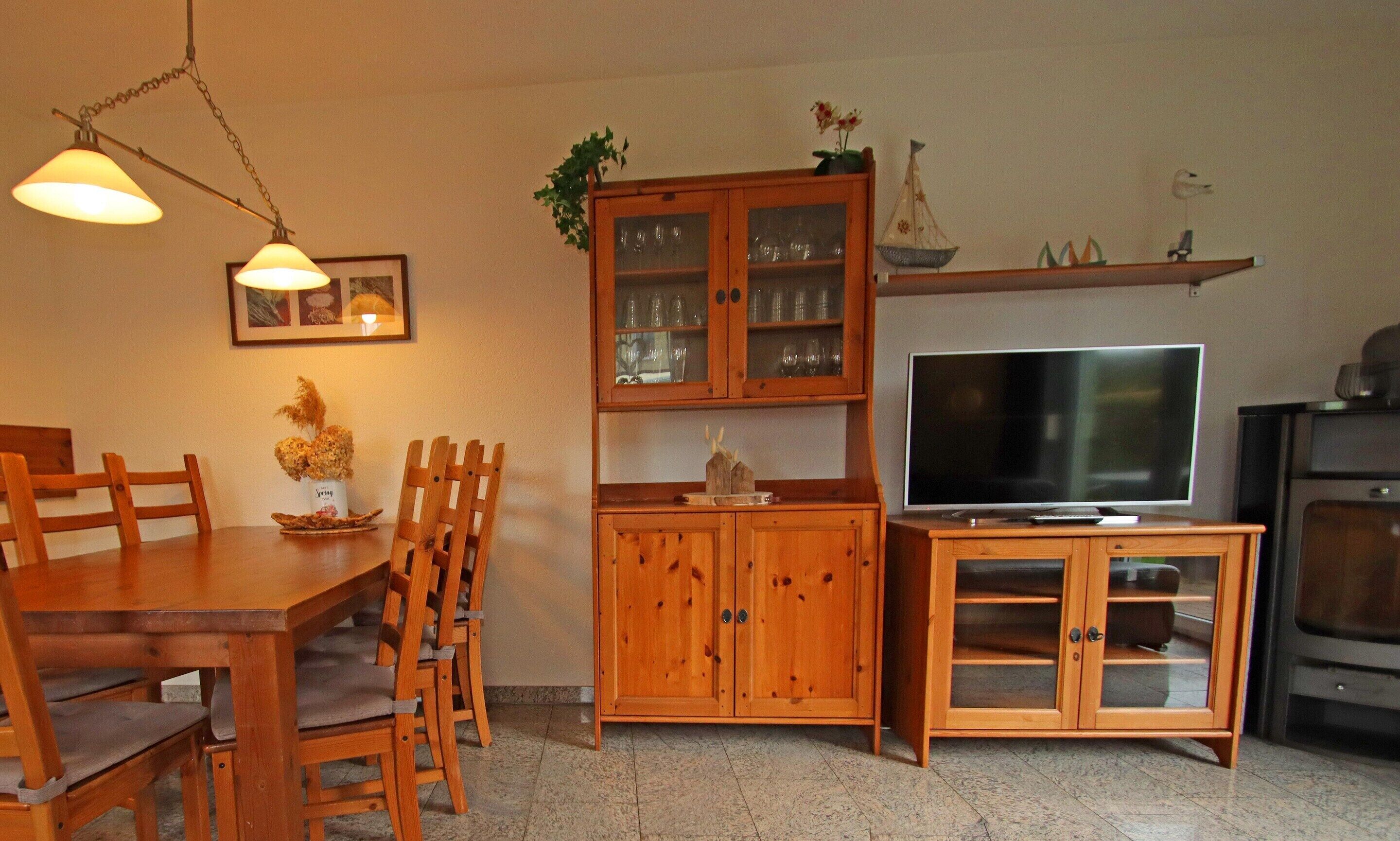 Una zona menjador amb una taula i cadires de fusta, un armari amb portes de vidre, una televisió sobre un suport de fusta i una estant amb objectes decoratius.