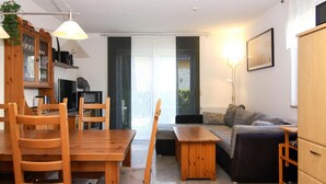 A cozy living room with a wooden dining table, chairs, a sofa, and a TV. There is a sliding glass door leading to a balcony. - Doppelhaushalfte, Zingst (Zingst)