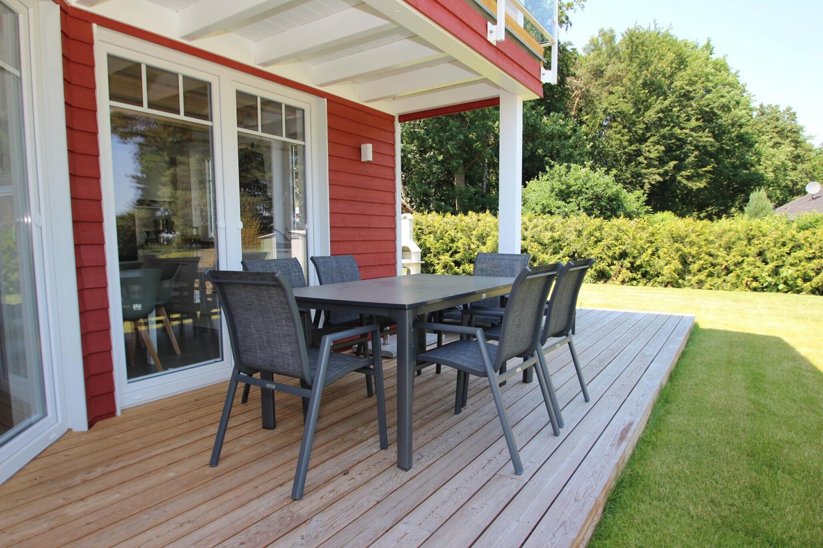 A covered patio with a table and chairs, a wooden deck, and a view of a green lawn and trees.