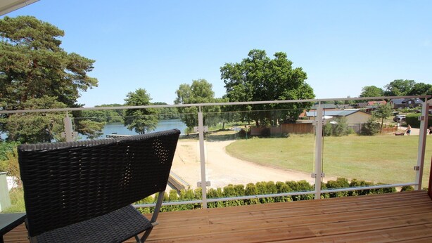 Eine Holzterrasse mit einem Korbstuhl, mit Blick auf einen See und eine grüne Fläche mit Bäumen und Häusern im Hintergrund.