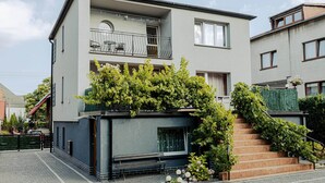 A modern two-story building with a balcony, a small garden area with plants, and a staircase leading to the entrance.