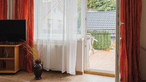 A room with a television on a wooden stand, a vase with dried plants, and a sliding glass door leading to an outdoor balcony with a tiled roof and greenery.