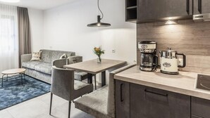 Cozinha moderna com cafeteira, chaleira e forno embutido. Há uma área de jantar com mesa e cadeiras, e um sofá ao fundo.