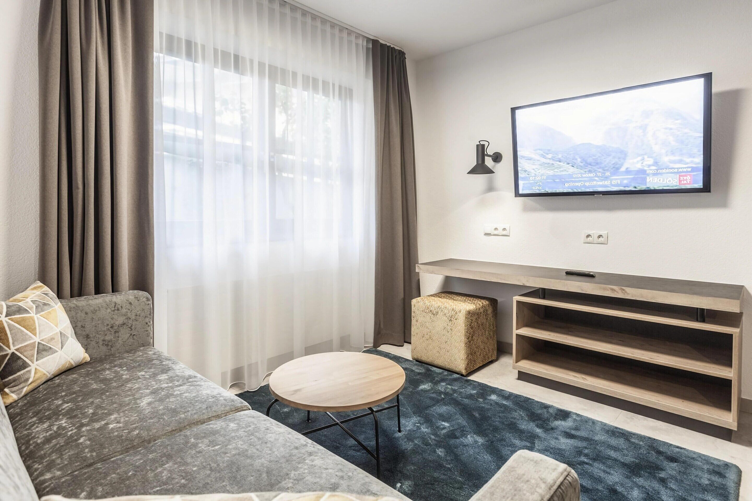 Een moderne woonkamer met een grijze bank, een ronde houten salontafel, een aan de muur bevestigde televisie en een houten tv-meubel.