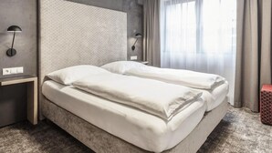 Quarto de hotel moderno com uma cama grande, duas mesas de cabeceira e uma janela ampla com cortinas.