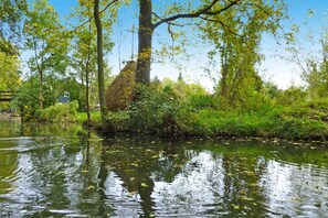 A serene riverside scene with a thatched-roof structure, lush greenery, and a calm water body reflecting the surroundings. - Apartments Spreeidyll, Schlepzig (Schlepzig)