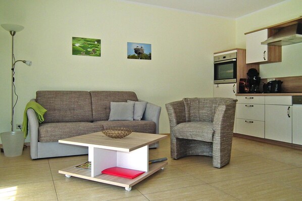 Sala de estar con sofá, sillón, mesa de centro, lámpara y una zona de cocina con armarios y electrodomésticos.