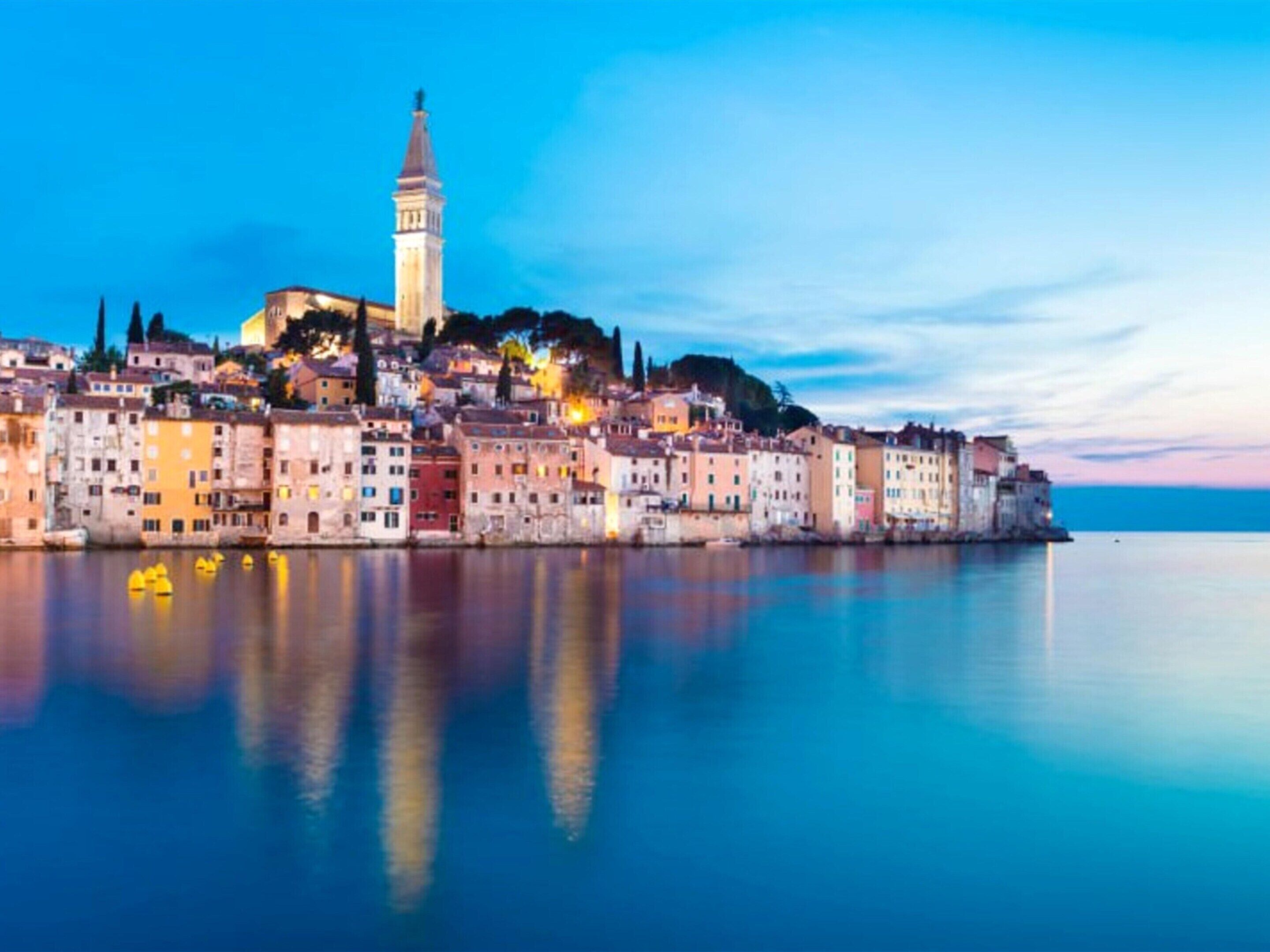 A coastal cityscape with a prominent tower, calm water reflecting buildings, and a clear sky.