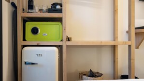 Ein kleiner Raum mit einem weißen TZS-Kühlschrank, einem grünen Safe und einem Holzregal mit diversen Gegenständen.