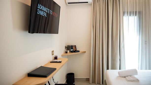 Ein modernes Hotelzimmer mit Flachbildfernseher, Klimaanlage, einem Schreibtisch mit einem Laptop, einem Bett mit einem zusammengerollten Handtuch und einem Fenster mit Vorhängen.