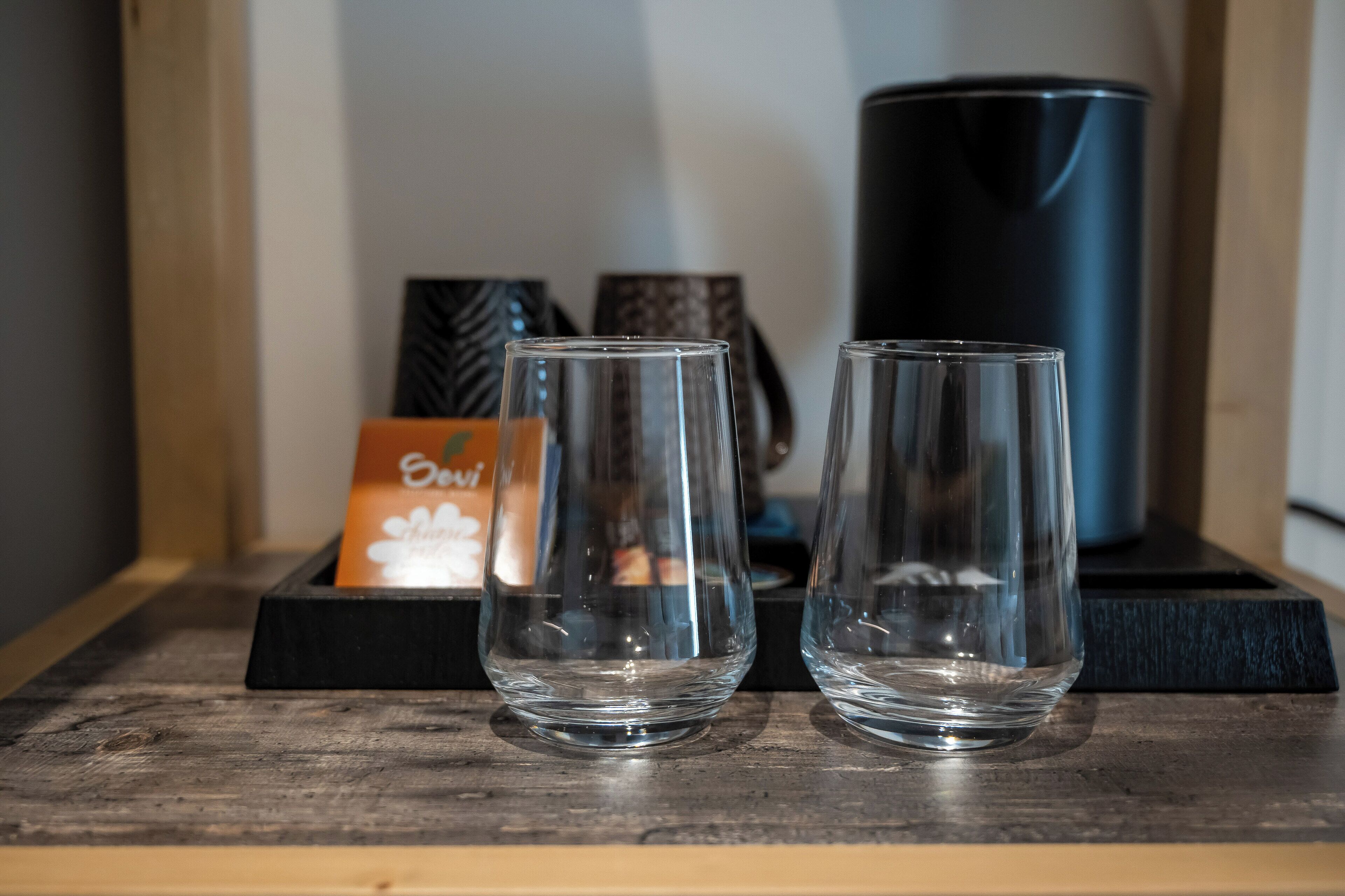 Two clear glasses on a wooden surface with a black tray, a blue container, and a card with the word "Ovi" in the background.