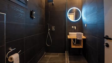 A modern bathroom with dark tiles, a glass shower enclosure, a round mirror, and a wooden vanity.