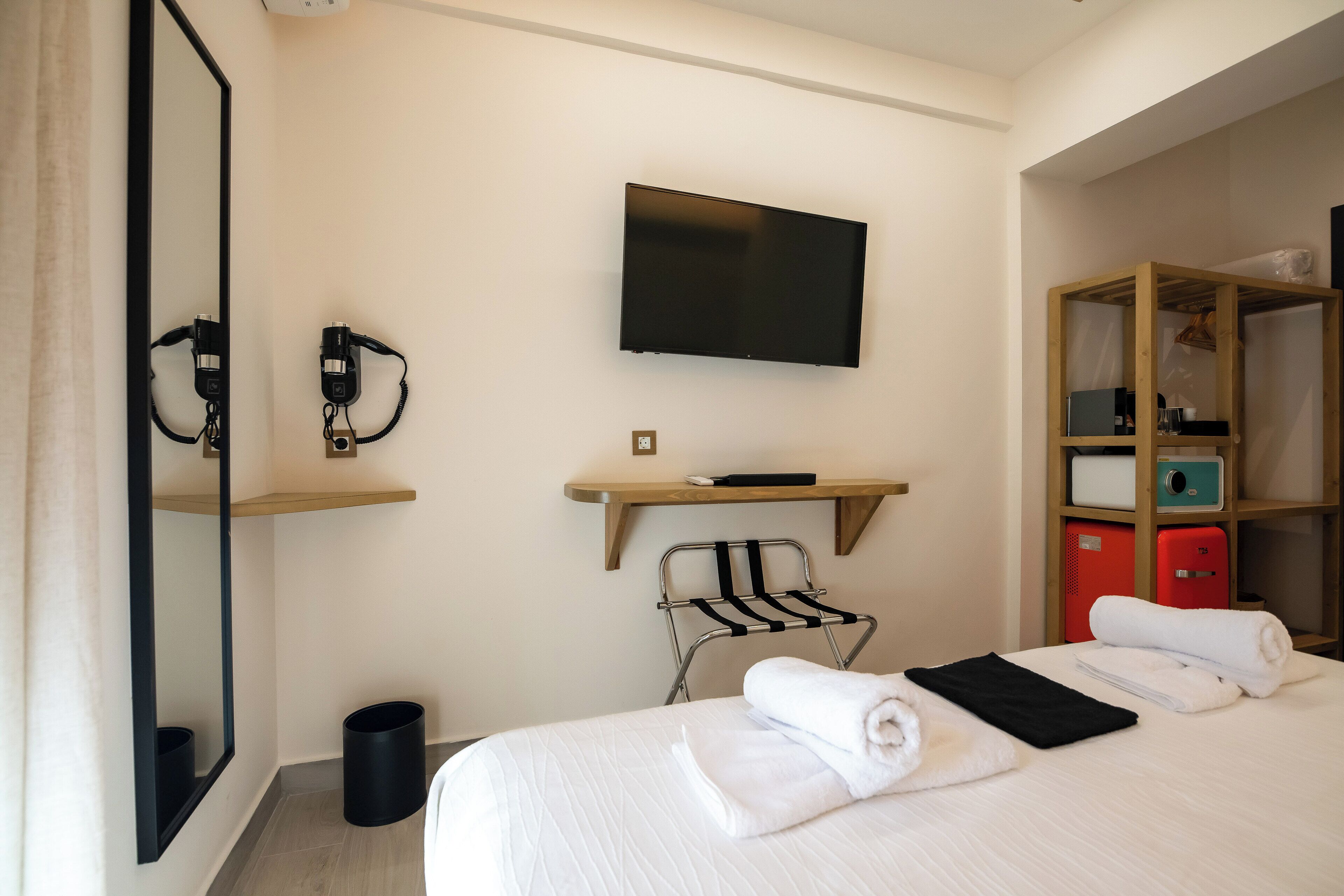 A hotel room with a bed, a TV mounted on the wall, a mirror, a towel rack, and a wooden shelf with a red suitcase.