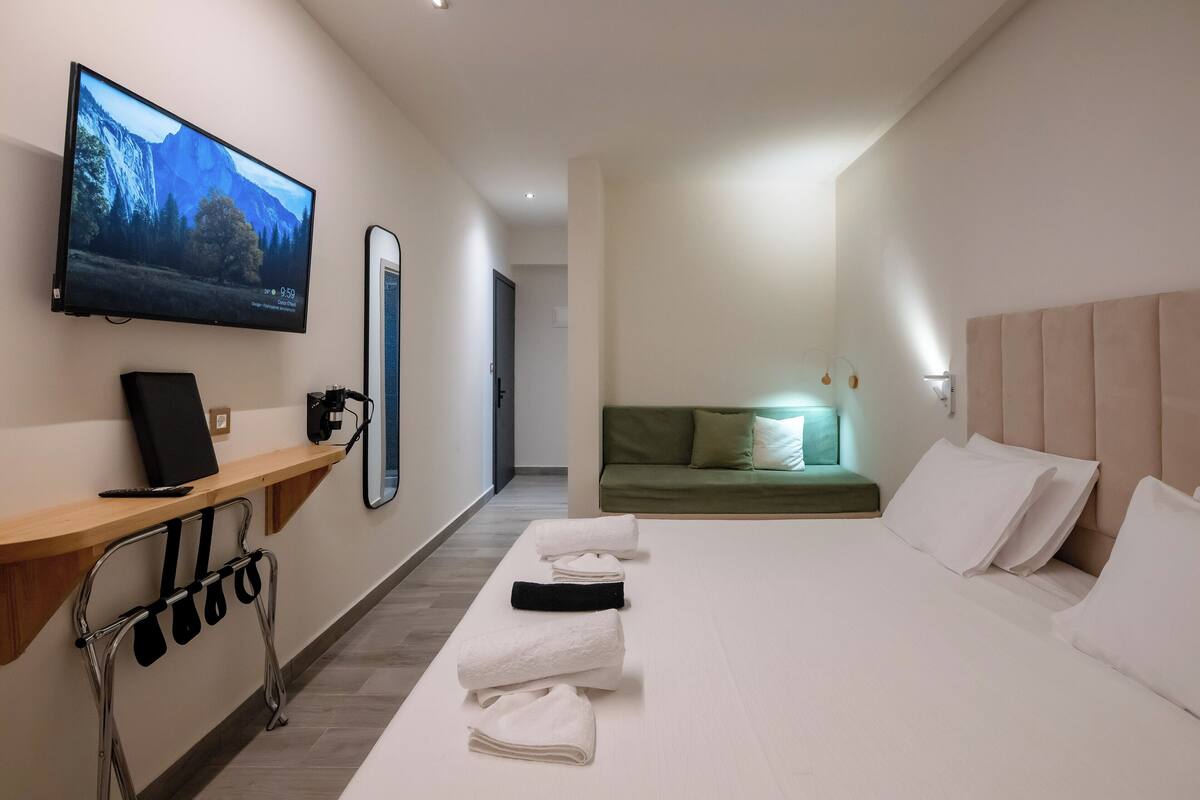 A hotel room with a large flat-screen TV mounted on the wall, a wooden desk with a chair, a bed with white linens, a green sofa, and a wall-mounted mirror.